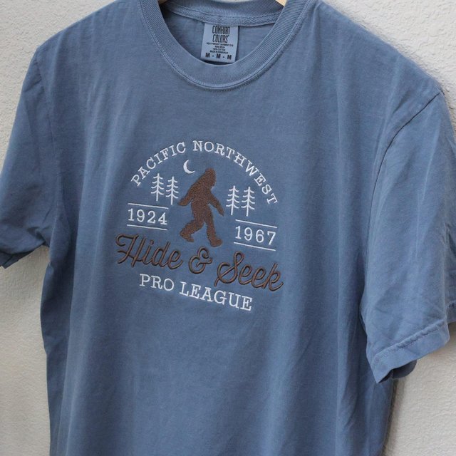 Close-up of an embroidered Pacific Northwest Hide and Seek Pro League design on a blue tee