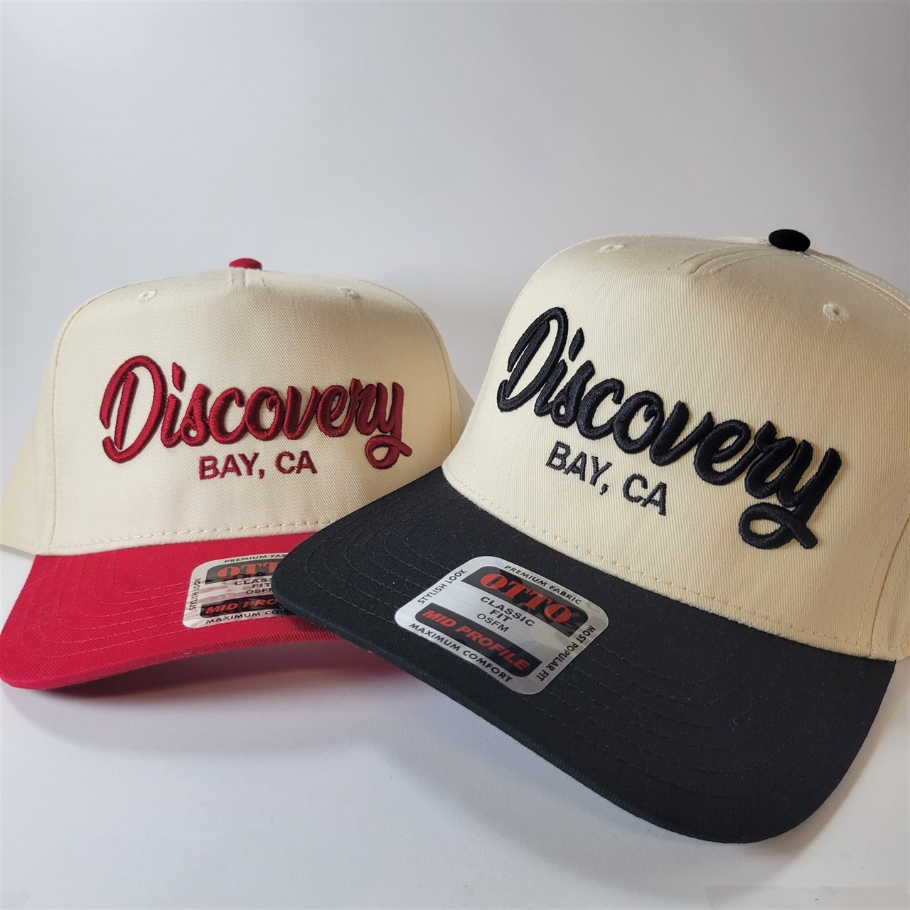 Two cream five-panel caps embroidered with Discovery Bay, CA in script, one with a red peak and one with a black peak